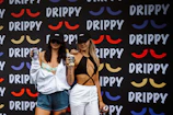 Two women wearing branded black caps hold beverage cans in front of a backdrop covered with colorful mustache-like designs and the word 'DRIPPY.' The woman on the left wears a light hoodie over a floral top and blue shorts, while the woman on the right wears a black top with white pants.