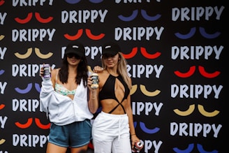 Two women wearing branded black caps hold beverage cans in front of a backdrop covered with colorful mustache-like designs and the word 'DRIPPY.' The woman on the left wears a light hoodie over a floral top and blue shorts, while the woman on the right wears a black top with white pants.