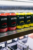 Rows of canned beverages displayed on a shelf, featuring branding that includes the words 'Drippy' and 'cannabis infused'. The cans are predominantly in red and yellow colors with black and white text. The background includes blurred shelves and packaging.