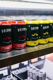 Rows of canned beverages displayed on a shelf, featuring branding that includes the words 'Drippy' and 'cannabis infused'. The cans are predominantly in red and yellow colors with black and white text. The background includes blurred shelves and packaging.