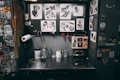 A tattoo workstation is cluttered with various items. The back wall displays numerous tattoo designs featuring hearts, daggers, skulls, and butterflies. The workbench holds ink bottles, paper towels, and various tattooing equipment. Stickers and artwork cover the surrounding surfaces, adding to the eclectic and artistic vibe.