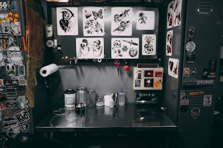A tattoo workstation is cluttered with various items. The back wall displays numerous tattoo designs featuring hearts, daggers, skulls, and butterflies. The workbench holds ink bottles, paper towels, and various tattooing equipment. Stickers and artwork cover the surrounding surfaces, adding to the eclectic and artistic vibe.