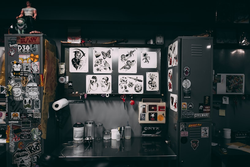 A cluttered workspace featuring a variety of tattoo designs, stickers, and art prints displayed on lockers and walls. The scene includes tools and supplies for tattooing, such as ink bottles, paper towels, and stencil paper. The area has a creative and informal vibe, with various art and pop culture references scattered throughout.