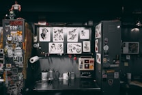 A cluttered workspace featuring a variety of tattoo designs, stickers, and art prints displayed on lockers and walls. The scene includes tools and supplies for tattooing, such as ink bottles, paper towels, and stencil paper. The area has a creative and informal vibe, with various art and pop culture references scattered throughout.