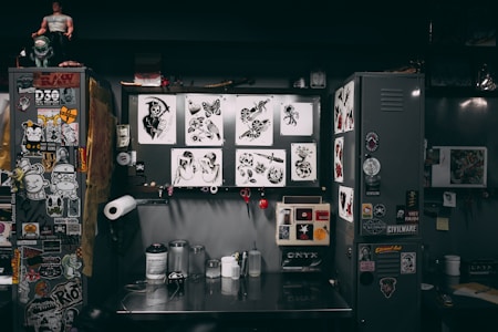 A cluttered workspace featuring a variety of tattoo designs, stickers, and art prints displayed on lockers and walls. The scene includes tools and supplies for tattooing, such as ink bottles, paper towels, and stencil paper. The area has a creative and informal vibe, with various art and pop culture references scattered throughout.