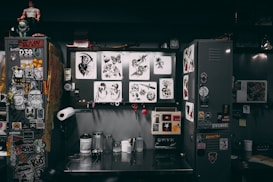 A cluttered workspace featuring a variety of tattoo designs, stickers, and art prints displayed on lockers and walls. The scene includes tools and supplies for tattooing, such as ink bottles, paper towels, and stencil paper. The area has a creative and informal vibe, with various art and pop culture references scattered throughout.