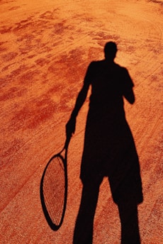 A shadow of a person holding a tennis racket is cast on a clay tennis court. The texture of the court is visible with its reddish-brown surface contrasting against the dark silhouette.