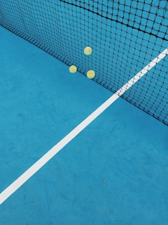 A vibrant blue tennis court with three tennis balls against a black net, intersected by a white line.