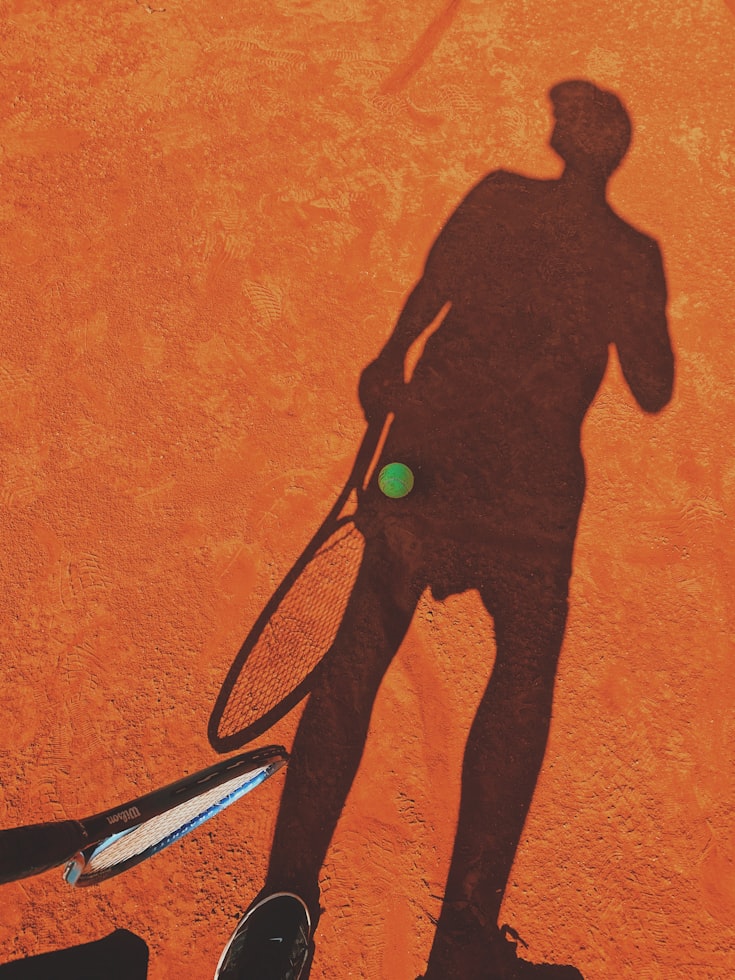 A solitary tennis player walking onto an empty court at sunset with a racket bag slung over the shoulder