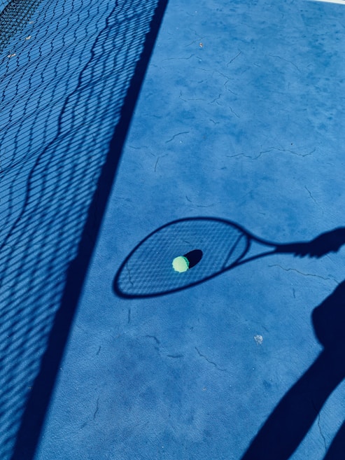 A tennis court with a vibrant blue surface and the shadow of a chain-link fence cast prominently across it. The shadow of a person holding a tennis racket is visible, with a tennis ball lying on the ground and framed by the racket's shadow.