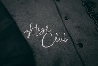 A close-up view of a grey fabric with the words 'High Club' embroidered in white cursive lettering. The fabric has a textured appearance, and there are black buttons visible on the right side.