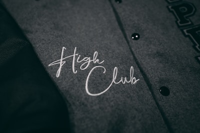 A close-up view of a grey fabric with the words 'High Club' embroidered in white cursive lettering. The fabric has a textured appearance, and there are black buttons visible on the right side.