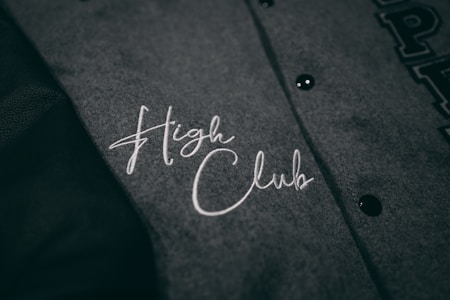 A close-up view of a grey fabric with the words 'High Club' embroidered in white cursive lettering. The fabric has a textured appearance, and there are black buttons visible on the right side.