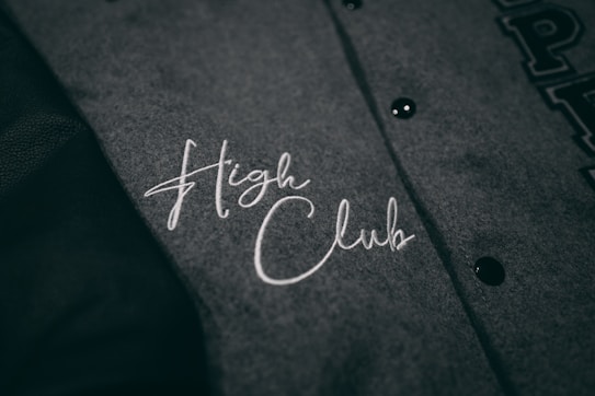 A close-up view of a grey fabric with the words 'High Club' embroidered in white cursive lettering. The fabric has a textured appearance, and there are black buttons visible on the right side.