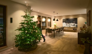 A person is decorating a Christmas tree in a warmly lit living room. The room has modern design elements, with a long dining table set with candles and ornaments. Elegant pendant lights hang above the table, and a large piece of abstract art adorns the wall. Soft, ambient lighting creates a cozy atmosphere.