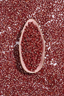 A large collection of red beans spread across a surface with a pink, elongated dish at the center filled with more beans.