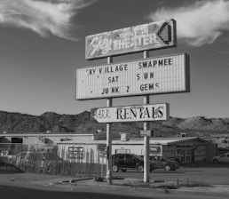 A vintage drive-in theater sign displays information for an event called Sky Village Swap Meet, taking place on Saturday and Sunday. The sign has a retro design with faded letters and stands against a backdrop of mountains and a few buildings. Several cars are parked near a fence, contributing to an old-fashioned, small-town atmosphere.