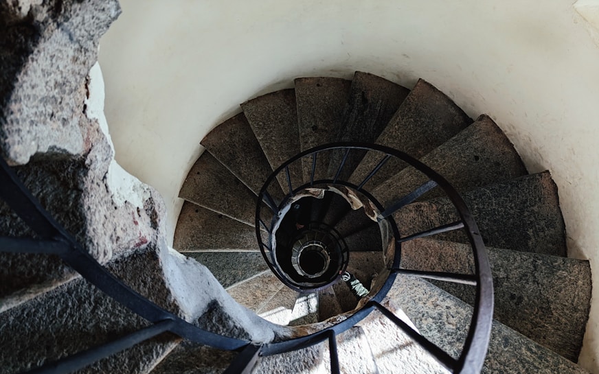 A spiral staircase made of stone and metal, viewed from above. The steps are rough textured and the railing is made of dark metal. The spiral pattern creates a dizzying effect in the confined space.