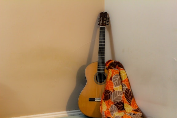 A classical guitar with a wooden finish leans against a corner where two walls meet. A colorful blanket with leaf patterns in shades of orange, yellow, brown, and cream is draped over the guitar.