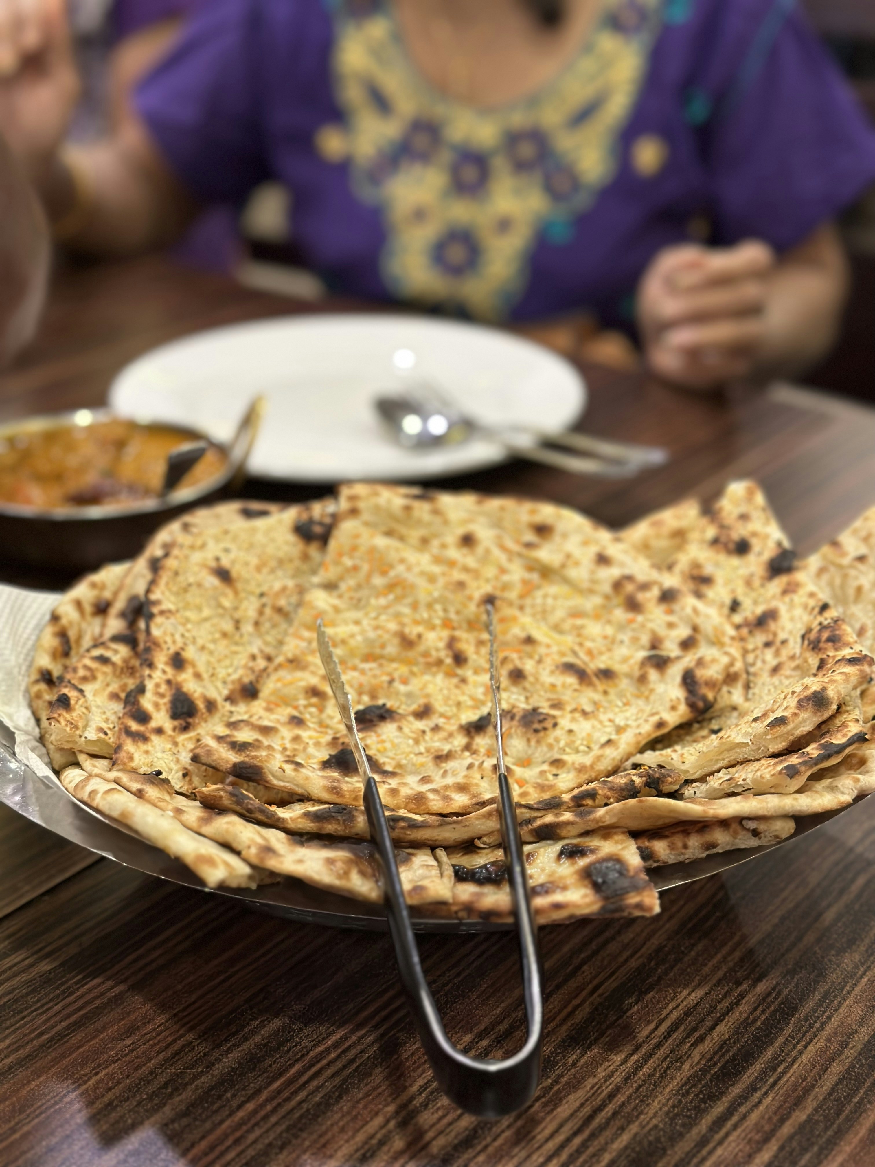 A stack of toasted flatbreads with steel tongs on a restaurant table, a curry in a steel bowl behind