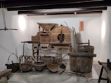 A display of traditional wooden farming tools and utensils arranged against a white wall. The assortment includes various baskets, a large wooden barrel, and a rustic machine with gears. The objects are predominantly made of wood and wicker, showcasing artisanal craftsmanship. A decorative orange sign with Chinese characters is mounted on the wall.