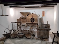 A display of traditional wooden farming tools and utensils arranged against a white wall. The assortment includes various baskets, a large wooden barrel, and a rustic machine with gears. The objects are predominantly made of wood and wicker, showcasing artisanal craftsmanship. A decorative orange sign with Chinese characters is mounted on the wall.