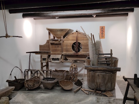 A display of traditional wooden farming tools and utensils arranged against a white wall. The assortment includes various baskets, a large wooden barrel, and a rustic machine with gears. The objects are predominantly made of wood and wicker, showcasing artisanal craftsmanship. A decorative orange sign with Chinese characters is mounted on the wall.
