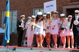 A group of young people is gathered on a stage, wearing white clothing and graduation caps. They are holding a sign with 'NA19b' and some wear medals around their necks. Some are holding roses and papers, suggesting a graduation ceremony. A blue and yellow flag is visible on the left, indicating a Swedish context.