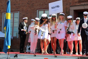 A group of young people is gathered on a stage, wearing white clothing and graduation caps. They are holding a sign with 'NA19b' and some wear medals around their necks. Some are holding roses and papers, suggesting a graduation ceremony. A blue and yellow flag is visible on the left, indicating a Swedish context.