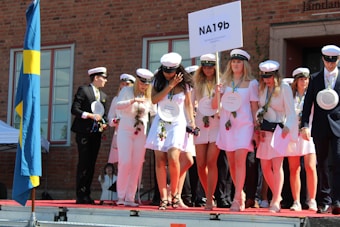 A group of young people is gathered on a stage, wearing white clothing and graduation caps. They are holding a sign with 'NA19b' and some wear medals around their necks. Some are holding roses and papers, suggesting a graduation ceremony. A blue and yellow flag is visible on the left, indicating a Swedish context.