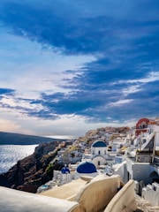 A stunning coastal town with iconic white-washed buildings and blue domed roofs perched on a hillside. The scene overlooks a vast, shimmering sea. The sky is partly cloudy, casting dynamic shadows and highlights across the landscape.