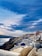 A stunning coastal town with iconic white-washed buildings and blue domed roofs perched on a hillside. The scene overlooks a vast, shimmering sea. The sky is partly cloudy, casting dynamic shadows and highlights across the landscape.