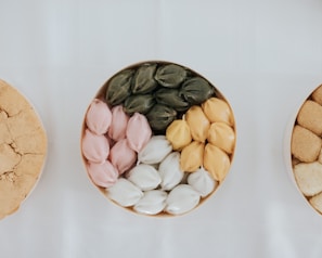 A beautifully arranged assortment of colorful sweets organized in a round container on a white background. The sweets are layered in four distinct colors: green, pink, white, and yellow, each set displaying a shiny, smooth texture.