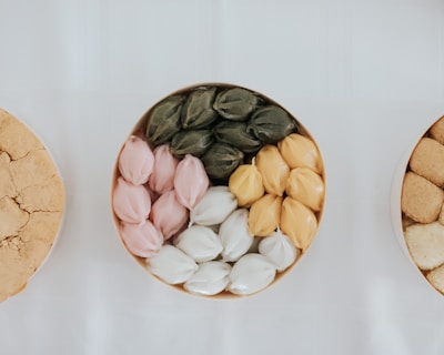 A beautifully arranged assortment of colorful sweets organized in a round container on a white background. The sweets are layered in four distinct colors: green, pink, white, and yellow, each set displaying a shiny, smooth texture.