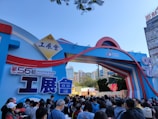 A large crowd of people gathered outside an entrance archway for the 56th Hong Kong Brands and Products Expo. The arch is decorated with colorful designs and Chinese characters, with buildings and trees visible in the background.