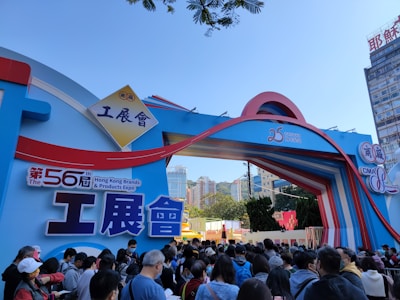 A large crowd of people gathered outside an entrance archway for the 56th Hong Kong Brands and Products Expo. The arch is decorated with colorful designs and Chinese characters, with buildings and trees visible in the background.