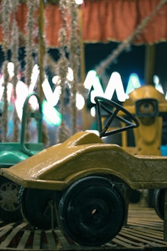 A vintage carousel with small ride-on cars, featuring a mustard-yellow car in the foreground and a green car partially visible. The backdrop is adorned with shimmering tinsel and festive lights that create a blurred bokeh effect, adding a nostalgic and whimsical atmosphere.
