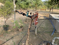 A garden setting featuring a series of pipes and valves connected to a water system. The setup includes various colored handles and a red cylindrical object likely functioning as a filter. Potted plants are scattered around the ground, some with leafy branches. The background is filled with lush green trees and a wooden bench, all under a clear sky.