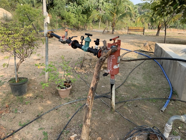 A garden setting featuring a series of pipes and valves connected to a water system. The setup includes various colored handles and a red cylindrical object likely functioning as a filter. Potted plants are scattered around the ground, some with leafy branches. The background is filled with lush green trees and a wooden bench, all under a clear sky.