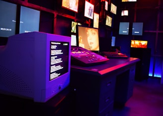 A dark room filled with multiple screens displaying various images and text. The setup includes a large CRT monitor showing text, a central monitor with a close-up image of a person's face, and several smaller screens on the surrounding walls. The lighting in the room is dim with neon blue and red hues, creating a moody and atmospheric ambiance.