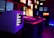 A dark room filled with multiple screens displaying various images and text. The setup includes a large CRT monitor showing text, a central monitor with a close-up image of a person's face, and several smaller screens on the surrounding walls. The lighting in the room is dim with neon blue and red hues, creating a moody and atmospheric ambiance.