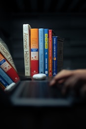 A row of self-help and business books stands upright on a table, adjacent to a laptop and a hand typing. The books have colorful covers, including titles like 'Atomic Habits' and 'The 4-Hour Work Week.' The setting appears dimly lit with a focused spotlight on the books, creating a cozy, studious ambiance.