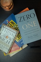 Several books are stacked on a dark surface, including 'Zero to One' by Peter Thiel and 'Think and Grow Rich' by Napoleon Hill. A pen lies next to the books and a lit candle in a copper holder is positioned nearby.