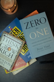 Several books are stacked on a dark surface, including 'Zero to One' by Peter Thiel and 'Think and Grow Rich' by Napoleon Hill. A pen lies next to the books and a lit candle in a copper holder is positioned nearby.