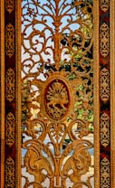 An ornate wrought iron gate with intricate patterns and designs, featuring a central emblem of a lion with a crown. The background reveals a glimpse of a garden and blue sky seen through the gate.