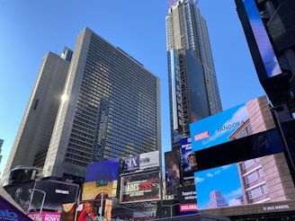 Tall skyscrapers dominate the skyline, featuring numerous large digital billboards displaying advertisements, including those for Marvel and Pandora. The scene conveys a bustling urban environment with bright, vivid colors against a clear blue sky.