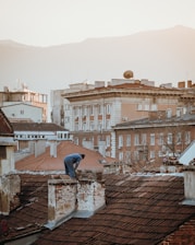 A person is working on the roof of an old building with red tiles, surrounded by other buildings of various architectural styles. The backdrop features mountains under a hazy sky. The image has a warm, soft light.