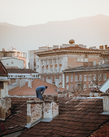 A person is working on the roof of an old building with red tiles, surrounded by other buildings of various architectural styles. The backdrop features mountains under a hazy sky. The image has a warm, soft light.
