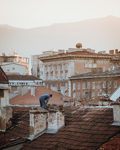 A person is working on the roof of an old building with red tiles, surrounded by other buildings of various architectural styles. The backdrop features mountains under a hazy sky. The image has a warm, soft light.