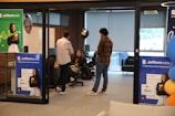A modern office setting features several people engaging in conversation. Posters advertising Jotform SIGN are prominently displayed on the walls and partitions. The space includes brick walls and large windows, with some natural light coming through. Balloons are in the background, adding a celebratory or casual atmosphere. The individuals are casually dressed, lending a relaxed tone to the environment.